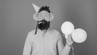 Kutlama kavramı. Sakal ve bıyık sakin yüz darbeler içine parti boynuz, mavi arka plan üzerinde olan adam. Hippi dev güneş gözlüğü Doğum günü kutluyor. Hava balonları ile parti şapkalı adamı kutluyor