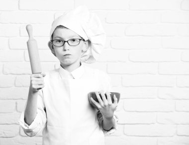 Genç çocuk küçük sevimli aşçı Şef beyaz üniforma ve şapka oklava tuğla duvar arka plan üzerinde kırmızı bir kase tutarak gözlüklü kızgın yüzündeki kopyalamak girintili