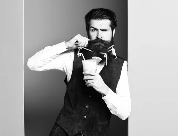 handsome bearded man with long beard and mustache has stylish hair on serious face holding glass of alcoholic beverage in vintage suede leather waistcoat on blue green studio background, copy spac