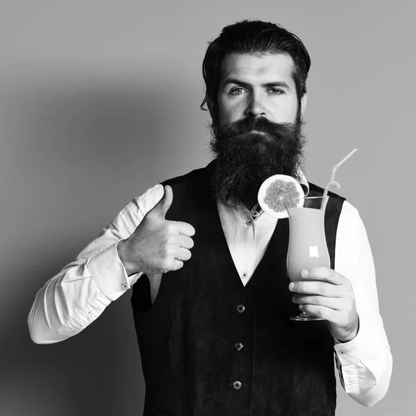 handsome bearded man with long beard and mustache has stylish hair on serious face holding glass of alcoholic cocktail in vintage suede leather waistcoat on blue studio background