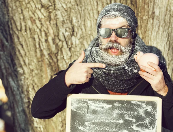 Upphetsad skäggig man, hipster, med skägg och mustasch i svarta solglasögon täckt med vit frost pekar på svarta tavlan och gris, spargris, vinterdag på naturlig bakgrund, kopiera spac — Stockfoto