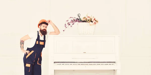 Man with beard, worker in overalls and helmet lean on piano, white background. Courier delivers furniture in case of move out, relocation. Loader moves piano instrument. Delivery service concept