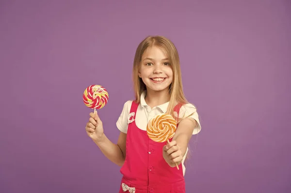 Essen und Dessert. Mädchen lächeln mit Lutschern auf violettem Hintergrund. glückliches Kind mit wirbelnden Karamellen auf lila Hintergrund. Ein kleines Kind lächelt mit Bonbons auf Stöcken. köstlich süß. Ernährung und Diät — Stockfoto
