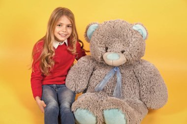 Gri yumuşak oyuncakla çocuk gülüşü. Turuncu arka planda büyük bir oyuncak ayıyla oturan kız. Hayvan oyuncaklı çocuk, hediye ve hediye. Tatil, doğum günü, yıldönümü kutlaması. Mutlu çocukluk kavramı.