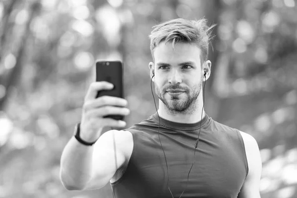 Sport gadget concept. Athlete mobile phone set up playlist before runnig. Man athlete busy face setting up smartphone app, nature background. Sportsman training with pedometer and earphones.