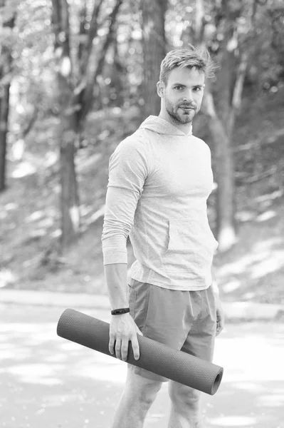 Practicing yoga. Sportsman carries yoga mat for outdoor practicing. Outdoor yoga concept. Man athlete on smiling face carries mat, going to stretching, nature background. Athlete with fitness tracker