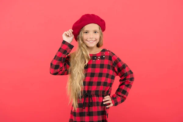 Kid söta lilla flicka med långa blonda hår poserar i basker hatt och rutig klänning röd bakgrund. Fashionabla beret tillbehör för kvinna. Beret stilinspiration. Bära basker som mode flicka — Stockfoto