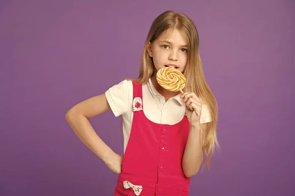 O miúdo come chupa-chupa no fundo violeta. Menina com caramelo redemoinho. Criança comendo doces em pau no fundo roxo. Comida e sobremesa. Dieta e dieta. Beijo gostoso — Fotografia de Stock