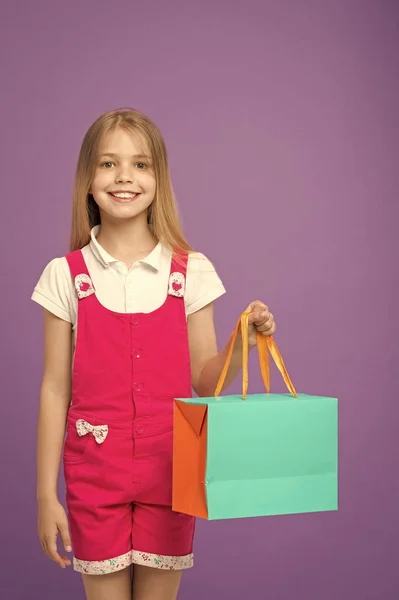 Małe dziecko z torbą na zakupy na fioletowym tle. Szczęśliwa dziewczyna uśmiech z worka papierowego. Dzieciak shopper w kombinezon mody. Święta i uroczystości. Zadowolony z jej zakupy. sprzedaż i czarny piątek — Zdjęcie stockowe