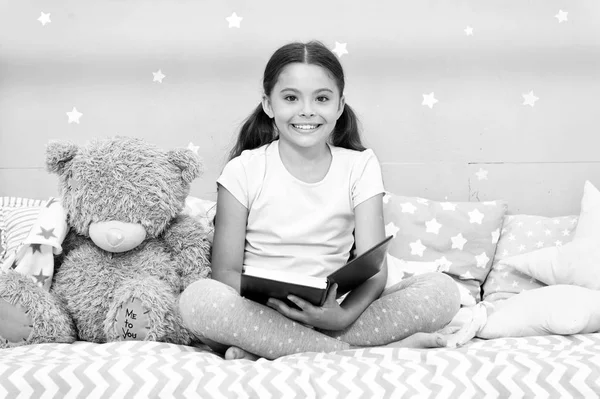 Leia conto de fadas antes de ir para a cama. Menina criança sentar cama com ursinho de pelúcia ler livro. O miúdo prepara-se para ir para a cama. Tempo agradável no quarto acolhedor. Menina miúdo longo cabelo bonito pijama relaxar e ler livro para carregar brinquedo — Fotografia de Stock