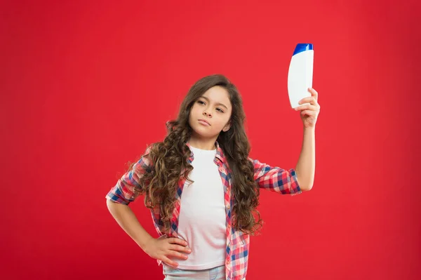 Chica pelo largo rizado celebrar botella de champú. Niña sobre fondo rojo. concepto de salón de peluquería. Champú para el cabello rizado. El bálsamo acondicionador nutre el cabello dañado. La mejor opción. Remedios para el cabello rizado — Foto de Stock