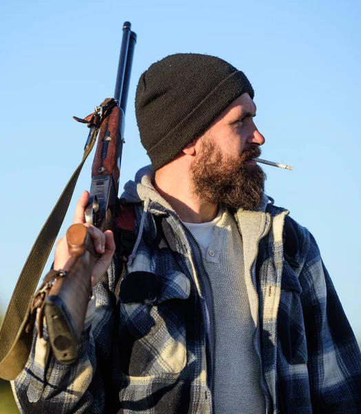 Hunting masculine hobby concept. Man brutal bearded guy gamekeeper blue sky background. Hunter with rifle gun close up. Guy bearded hunter spend leisure hunting and smoking. Brutality and masculinity