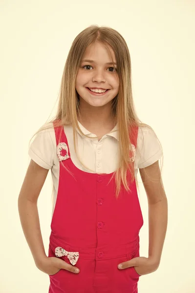 Barnet ler med långa blonda hår. Liten flicka leende i rosa jumpsuit isolerade på vitt. Kid modell i fashionabla övergripande. Stil och trend. Lycklig barndom och barnomsorg — Stockfoto