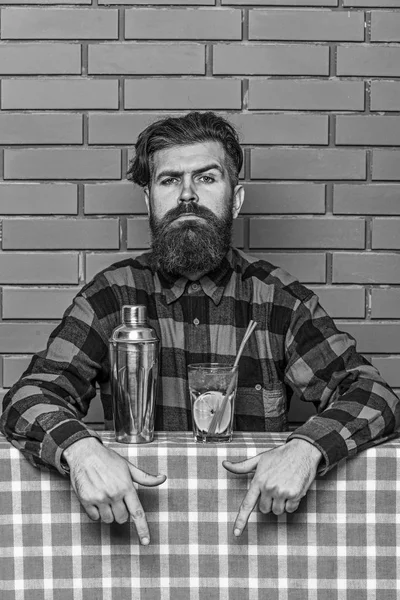 Bar menu concept. Bartender recommends to try beverage. Barman with beard on strict face pointing down with index finger. Man in checkered shirt near shaker and cocktail, brick wall background