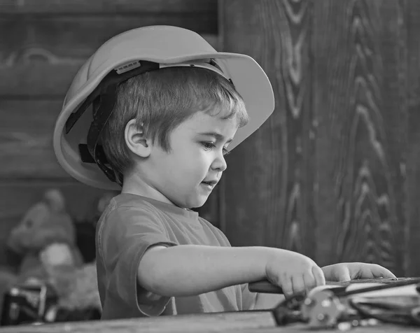Ребёнок играет за столом с инструментами. Концепция ручной работы. Ребенок в шлеме мило играть в качестве строителя или ремонтника, ремонт или ручной работы. Тоддлер на занятом лице играет дома в мастерской — стоковое фото