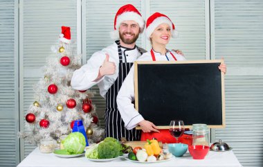 Erkek ve kadın şef santa şapka Noel ağacı yakınındaki blackboard kopya alanı tutun. Noel yemeği için çift onay listesi malzemeler. Noel tarifi kavramı. Bizim aile için yemek listesi. Christmas yemek pişirme