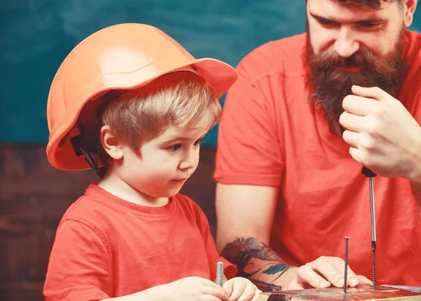 Мальчик, ребенок, занятый в защитном шлеме, учится пользоваться отвёрткой с папой. Отец, родитель с бородой учит маленького сына использовать отвёртку. Концепция ремонта и мастерской — стоковое фото