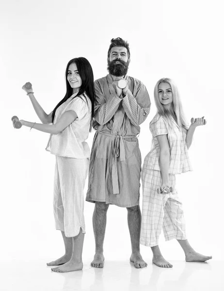 Bearded man with sporty ladies. Trainer, macho with attractive girls do morning exercises. Fitness morning concept. Company of friends with dumbbells in domestic clothes, isolated on white background