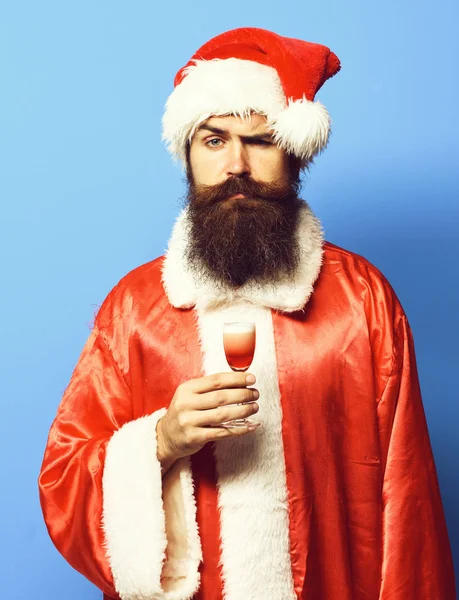 Красивий бородатий Санта Клаус чоловік з довгою бородою на серйозному обличчі, тримає келих алкоголю, знятий у червоному різдвяному або різдвяному светрі та новорічному капелюсі на синьому студійному фоні — стокове фото