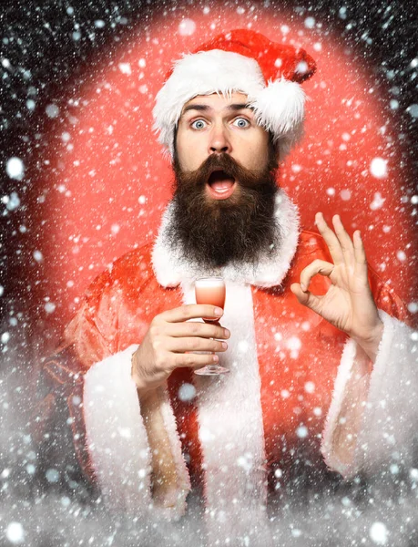Szép szakállas Mikulás ember, hosszú szakállal, a surprisedface holding üveg alkoholos lövés a karácsony vagy a karácsony kutyaruhák és újévi hat, és bemutatja rendben van a piros stúdió háttere — Stock Fotó