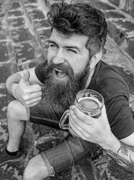 Man with beard and mustache holds glass with beer and shows thumb up, stone stairs background. Hipster on cheerful face drinking draft beer outdoor. Macho having rest with beer. Friday relax concept — Stock Photo, Image