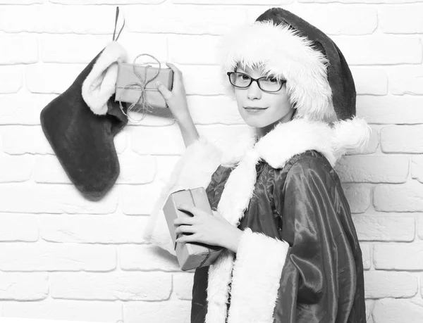 Kraft kağıt beyaz tuğla duvar arka plan üzerinde kırmızı kazak ve yeni yıl şapkalı dekoratif Noel veya Noel çorap veya önyükleme hediyeler holding ile gözlüklü genç sevimli Noel Baba çocuk sarılı — Stok fotoğraf