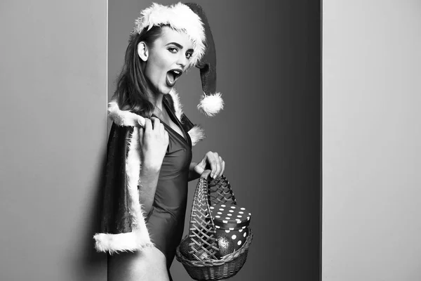 Πολύ χαριτωμένα σέξι κορίτσι santa ή χαμογελώντας ξανθιά γυναίκα με μόδα μπλε χείλη στο νέο έτος μαγιό και καπέλο κρατά διακοσμητικό καλάθι Χριστούγεννα ή Χριστούγεννα με πολύχρωμες μπάλες σε φόντο κόκκινο πράσινο στούντιο — Φωτογραφία Αρχείου
