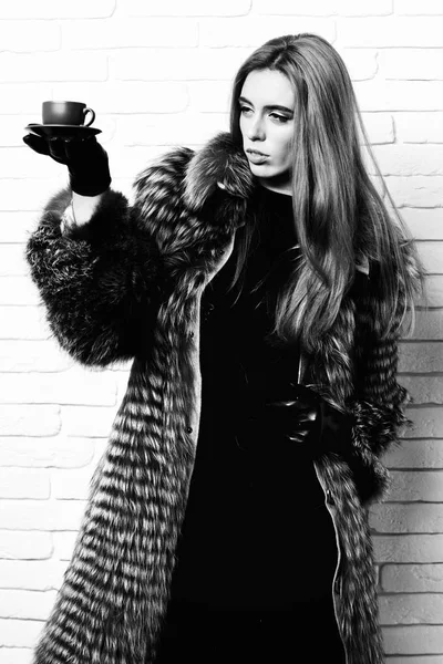 Молода сексуальна досить багата жінка з красивим довгим світлим волоссям на талії пальто сірого хутра з чорними шкіряними рукавичками, що тримає коричневу керамічну чашку кави на цегляному фоні студії — стокове фото