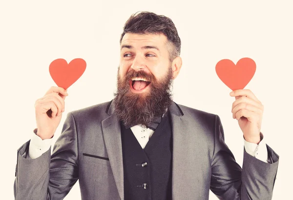 Hipster usa traje formal. Hombre feliz con barba larga y cara sonriente. El hombre barbudo sonríe y sostiene los corazones rojos. Citas, amor, romance, símbolo, moda, estilo, emociones, sentimientos, concepto de coqueteo — Foto de Stock