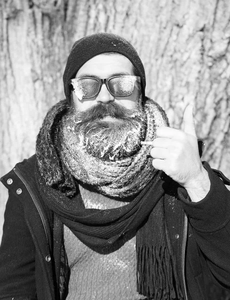 Beau barbu homme, hipster, avec barbe et moustache dans des lunettes de soleil noires recouvert de givre blanc fume cigarette sur le jour d'hiver à l'extérieur sur fond naturel, tient le pouce u — Photo