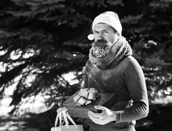 Hombre guapo en sombrero de Santa Claus, hipster barbudo con barba y bigote cubierto con escarcha blanca, sostiene caja de regalo roja, bolsas, regalos en el soleado día de invierno al aire libre en el fondo natural —  Fotos de Stock