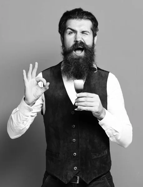 Schöner bärtiger Mann mit langem Bart und Schnurrbart auf lustigem Gesicht mit einem Glas alkoholischen Schnaps in einer Vintage Wildlederweste auf rotem Studiohintergrund — Stockfoto