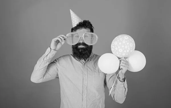 Hipster mit Riesensonnenbrille feiern Geburtstag. Mann mit Bart und Schnurrbart auf glücklichem Gesicht hält Bündel Luftballons, roter Hintergrund. Festkonzept. Mann mit Partyhut feiert Feiertag — Stockfoto