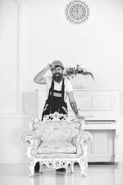 Movedor feliz em pé atrás da poltrona vintage com estampa florida. Barbudo cara em capacete laranja na frente do piano isolado no fundo branco — Fotografia de Stock