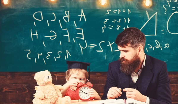 Učitel s vousem, otec učí malého syna v učebně, tabuli na pozadí. Chlapče dítě na klidnou tváří drží budík, zatímco učitel mluvit dítě. Individuální lekce koncept — Stock fotografie
