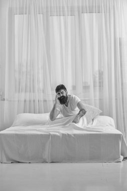 Akşamdan kalma kavramı. Adam sabah uyanma hayal kırıklığına acı yüzü. Tişörtlü adam yatak uyanık, beyaz perde arka plan üzerinde oturur. Akşamdan kalma maço sakal ve bıyık uğrar