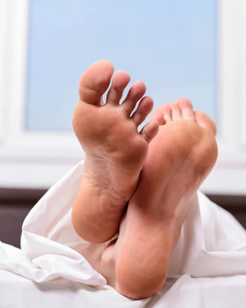 Feet appear out of blanket close up. Man sleeping on bed under blanket. Sleep alone. Healthy skin on foot. Size of foot. Male feet on bed in morning. Fresh and relaxed. Health and wellbeing concept