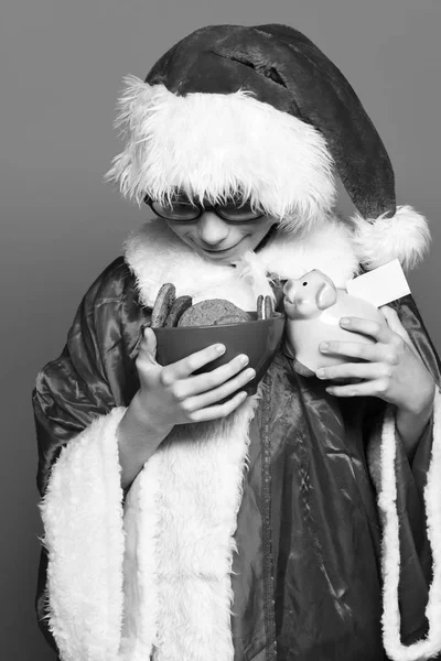 Kırmızı kazak ve yeni yıl Noel şapka pembe domuz domuz banka ve çikolata kalça kurabiye kase mavi stüdyo arka planda tutan genç Sevimli Noel baba çocuk ile gözlük — Stok fotoğraf