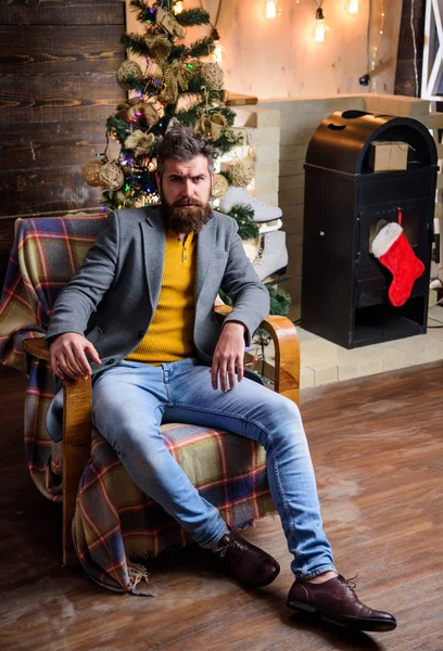 Man bearded hipster relax sit armchair near christmas tree. Feel like home. Man mature confident guy with beard relax at home. Warm and cozy christmas atmosphere at home. Home is best place