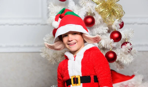 Traje de elfo de Navidad para niño. Fiesta de Navidad con disfraz de elfo. Ideas de árboles de Navidad para niños. Niño vestido como lindo elfo criatura mágica blanco orejas artificiales y sombrero rojo cerca del árbol de Navidad —  Fotos de Stock