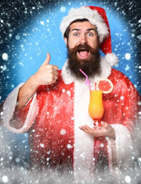 Schöner bärtiger Weihnachtsmann mit langem Bart auf lustigem Gesicht, der ein Glas alkoholfreien Cocktails in Weihnachts- oder Weihnachtspullover und Neujahrsmütze auf blauem Studiohintergrund hält — Stockfoto