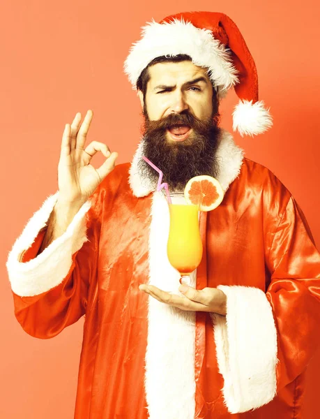 Schöner bärtiger Weihnachtsmann mit langem Bart auf glücklichem Gesicht, der ein Glas alkoholfreien Cocktails in Weihnachts- oder Weihnachtspullover und Neujahrsmütze auf rotem Studiohintergrund in der Hand hält — Stockfoto