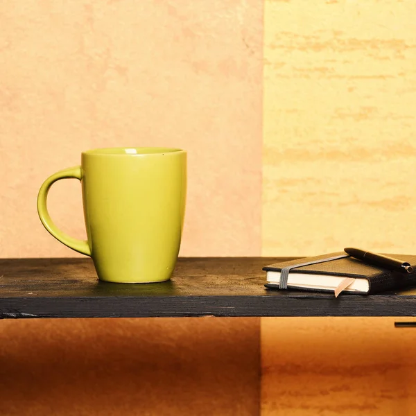 Cuaderno con marcador, pluma y taza verde en la mesa cerca de la pared . — Foto de Stock