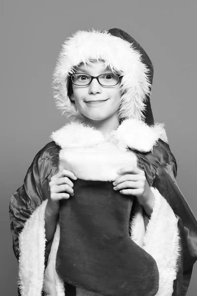 Joven lindo niño de Santa Claus con gafas en suéter rojo y sombrero de año nuevo sostiene la Navidad decorativa o medias de Navidad o bota sobre fondo de estudio azul —  Fotos de Stock
