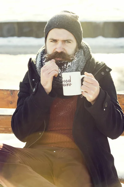 Rynka pannan man, skäggiga hipster med skägg och mustasch täckt med vit frost drycker från cup med god morgon texten sitter på träbänk på snöig vinter dag utomhus på naturliga bak — Stockfoto