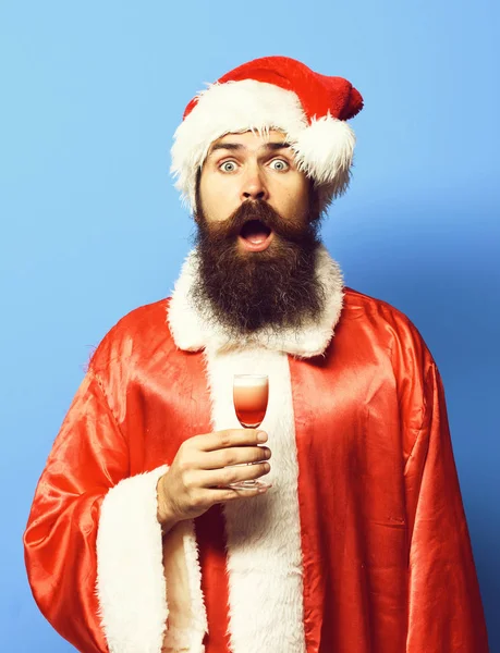 Guapo barbudo hombre de Santa Claus con barba larga en la cara sorprendida sosteniendo vaso de trago alcohólico en rojo navidad o suéter de Navidad y sombrero de año nuevo sobre fondo de estudio azul —  Fotos de Stock