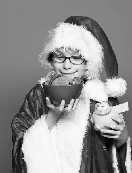 Jovem bonito santa claus menino com óculos em camisola vermelha e chapéu de Natal ano novo segurando rosa porquinho banco e chocolate biscoitos hip na tigela no fundo do estúdio azul — Fotografia de Stock