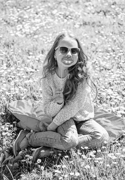 Fille s'assoit sur l'herbe à la prairie, fond vert. Enfant posant avec des lunettes en carton lèvres souriantes pour une séance photo au pré. Concept d'humour. Fille sur le visage joyeux passer des loisirs en plein air — Photo