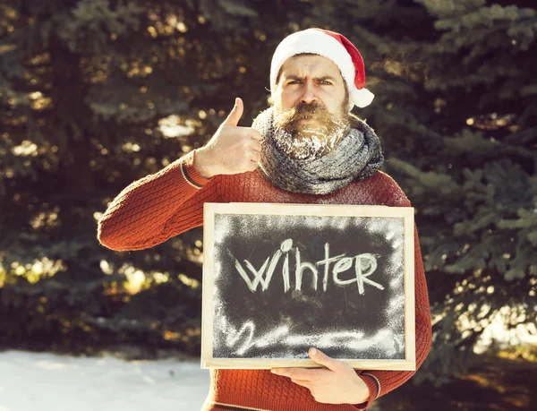 Verwirrter Mann mit Weihnachtsmann-Hut, bärtiger Hipster mit Bart und weißem Rauschebart, hält Daumen hoch, mit Winterwort auf schwarzem Brett sonniger Tag auf natürlichem Hintergrund — Stockfoto
