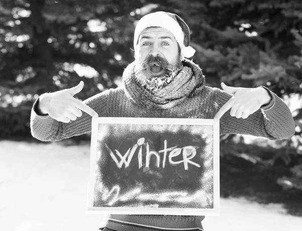 Homem alegre em chapéu de santa claus, hipster barbudo com barba e bigode coberto com geada branca, pontos em placa preta ou quadro com palavra de inverno no dia ensolarado ao ar livre em backgroun natural — Fotografia de Stock